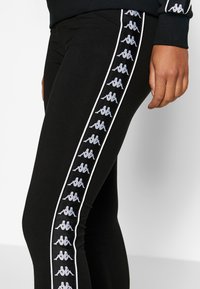 Czarne legginsy o dopasowanym kroju, z kontrastującym białym paskiem po boku i powtarzającymi się szarymi grafikami logo wzdłuż paska. Miękki, elastyczny materiał.