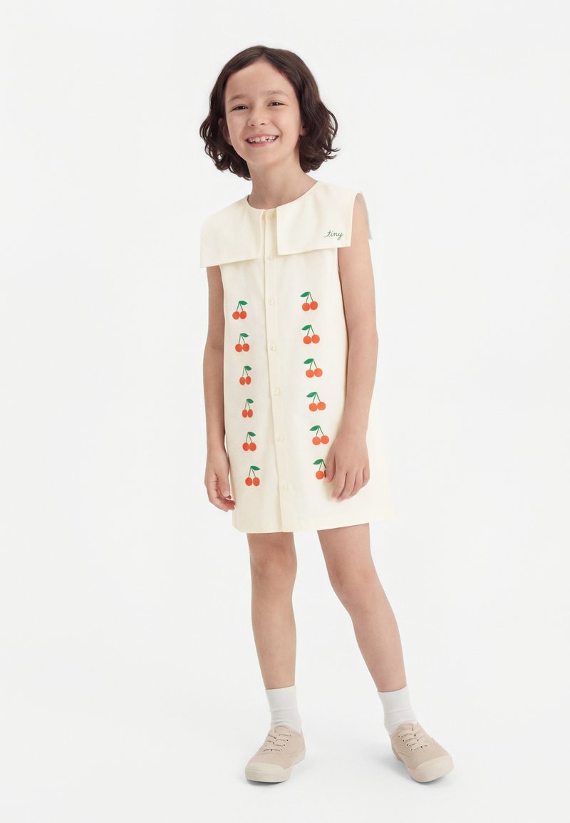 Χαμογελαστό παιδί που φοράει ένα αμάνικο κρεμ φόρεμα με μεγάλο γιακά και σχέδια με κερασάκια, σετ με λευκές κάλτσες και μπεζ αθλητικά παπούτσια.