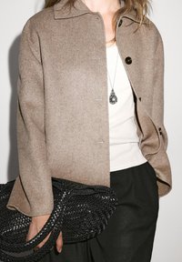 Brun uld-blandet jakke med krave, trykknaplukning og to synlige lommer. Sort vævet taske med tykke fletbåndshåndtag.