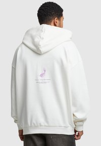 Sudadera blanca con una gran capucha y puños acanalados. La parte trasera presenta un gran gráfico en lavanda y texto en púrpura. Tejido suave, corte relajado.