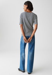 Femme debout avec les mains jointes derrière, portant une chemise rayée bleu marine et blanche, un jean bleu et des chaussures noires sur un fond uni.