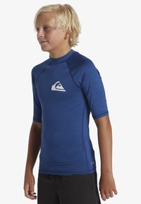 Blå rashguard med korta ärmar och slät textur, med en vit logotyp på bröstet och raglanärmar för ökad rörlighet. Svarta shorts.