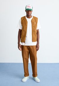 Man stands on blue floor against white wall wearing brown fleece vest, white t-shirt, brown pants, green and white checkered slip-on shoes, and green cap.