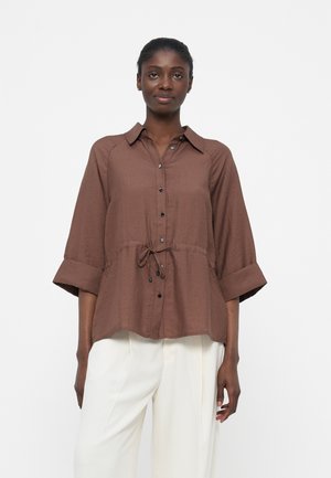 Ženska s temno poltjo in z lasmi zavezanim nazaj nosi rjavo srajco na gumbe z vrvico v pasu in zavihanimi rokavi, kombinirano z belimi hlačami.