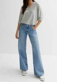 Maglia grigia a coste con scollatura a V, abbinata a jeans a gamba larga blu chiaro. Sneakers bianche visibili, che offrono uno stile casual.