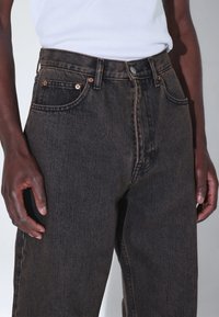 Mustat korkeavyötäröiset farkut, joissa kuparinväriset metalliosat, viisi taskua ja kontrastinen ompelu. Teksturoidun kankaan pinta on näkyvissä.