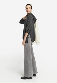 Dunkelgrauer Langarm-Hoodie mit seitlichen Schlitzen, kombiniert mit hellgrauen, weiten Jeans. Das Model trägt schwarze Schuhe und hält einen weißen Sweatshirt.