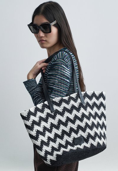 Γυναίκα που φορά μαύρα γυαλιά ηλίου και ριγέ τοπ, κρατά μια μεγάλη μαύρη και άσπρη τσάντα ώμου με ζιγκ ζαγκ σχέδιο.