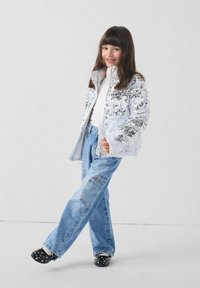 Chaqueta plateada con lentejuelas y cremallera, mangas blancas con puños, combinada con vaqueros holgados azul claro y zuecos negros con lunares.