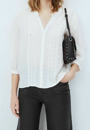 Femme portant un chemisier blanc brodé et un pantalon noir, tenant un sac à bandoulière noir clouté, une main dans la poche.