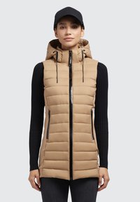 Gilet imbottito beige con cappuccio e chiusura zip, caratterizzato da cerniere laterali nere e un motivo trapuntato. Indossato sopra una maglietta nera a maniche lunghe a costine.
