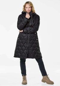 Schwarze wattierte Steppjacke mit Kapuze, ausgestattet mit einem Reißverschluss, Druckknöpfen und elastischen Kordeln. Kombiniert mit dunklen Jeans und braunen Stiefeln.