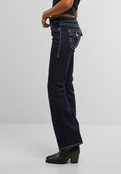 Jean en denim bleu foncé avec des coutures blanches contrastées et un design évasé. Présente des détails uniques sur les poches arrière et une taille mi-haute.