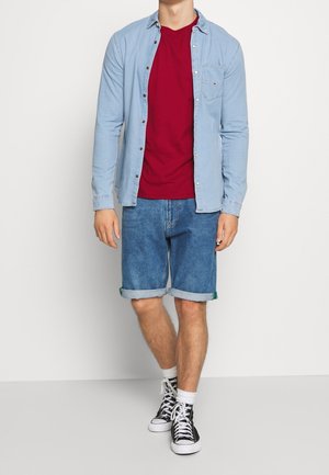 Jasnoniebieska koszula na guziki noszona na czerwonym T-shircie, w zestawie z dżinsowymi szortami. Czarne trampki z białą podeszwą oraz podwinięte mankiety szortów.