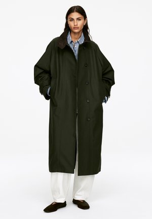 Long, dark green trench coat with a brown collar, featuring button closure and side pockets, worn over a striped shirt and white pants.
