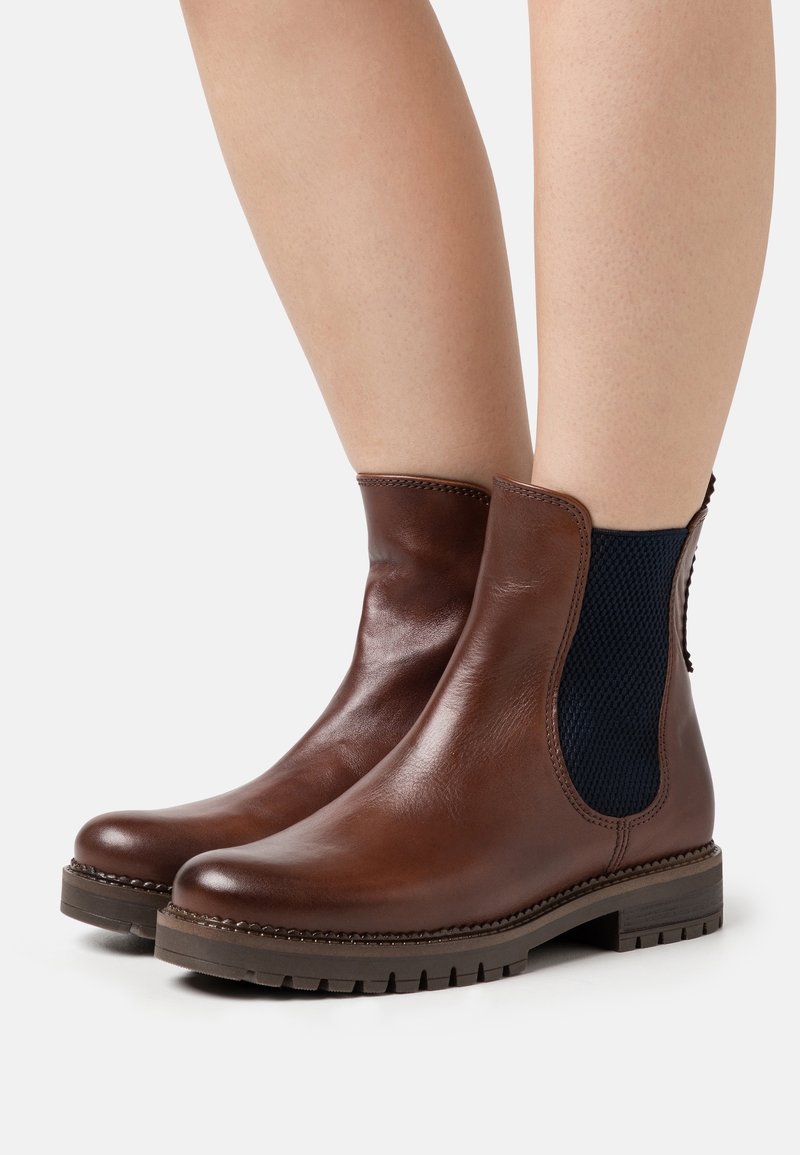Bottes courtes en cuir marron avec des panneaux élastiques noirs sur les côtés et des semelles en caoutchouc épaisses, portées sur des jambes nues, présentées sur un fond blanc.