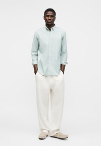 Mann som står med hendene i lommer, iført en lyseblå skjorte med knapper, hvite vide bukser og beige sko mot en hvit bakgrunn.