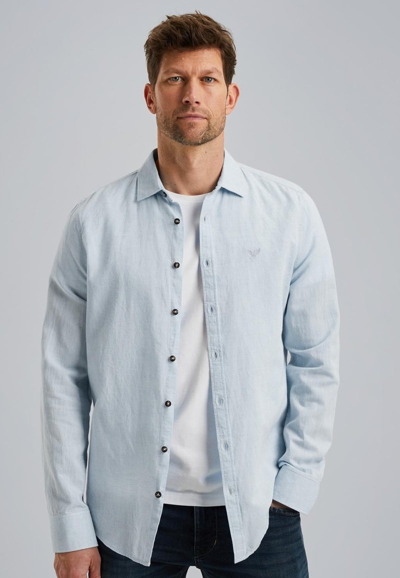 Camisa de manga larga azul claro con cuello tipo button-down, que presenta botones oscuros y un logo sutil en el pecho, llevada sobre una camiseta blanca.