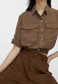 Chemise marron courte avec deux poches poitrine, boutons décoratifs et manches retroussées, associée à un pantalon marron taille haute avec grandes poches latérales.