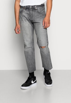 Mand iført lysegrå slidte jeans med hul ved knæet, sorte sneakers med hvide såler og en hvid t-shirt, stående på en grå gulv.