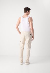 Pantalones cargo beige de algodón, con bolsillos laterales y un corte holgado. Se llevan con una camiseta sin mangas blanca y zapatos blancos.