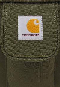 Olivenfarvet stofpose med en foldet klap og en hvid etiket, der viser et gult logo og "carhartt" i maroon tekst. Tekstureret overflade.