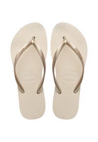 Beige flip-flops dengan sol berpola, menampilkan tali karet berwarna emas dan logo pada tali. Desain yang ringan dan minimalis.