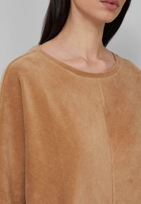 Vrouw in een ribbelige, tan kleurige corduroy top met een ronde halslijn en lang, donker haar zichtbaar tegen een effen lichtgrijze achtergrond.