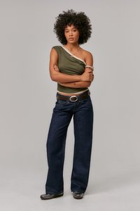 Top verde oliva a spalla unica con accento neutro, abbinato a jeans a gamba larga blu scuro e una cintura marrone. La modella sta in posizione con le braccia incrociate.