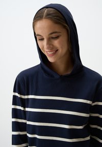 Felpa con cappuccio blu navy con strisce orizzontali bianche. Presenta una texture morbida e una vestibilità comoda, enfatizzando il design casual.