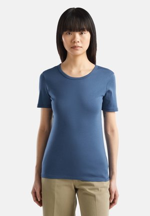 Giovane donna con capelli neri lisci che indossa una maglietta blu attillata con maniche corte e pantaloni beige su uno sfondo uniforme.