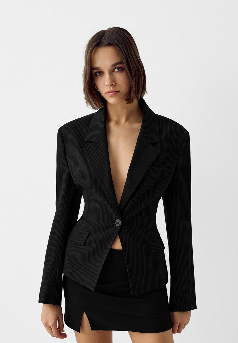 Joven mujer con cabello castaño hasta los hombros, vestida con un blazer negro de rayas y una falda mini negra con una abertura frontal.