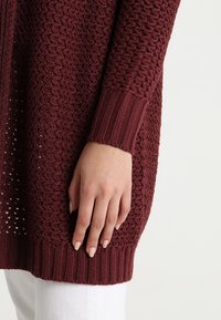 Burgundy stickad tröja med öppet vävmönster, ribbade manschetter och texturerad kropp. Mjuk material, avslappnad passform och detaljerad sömnad.