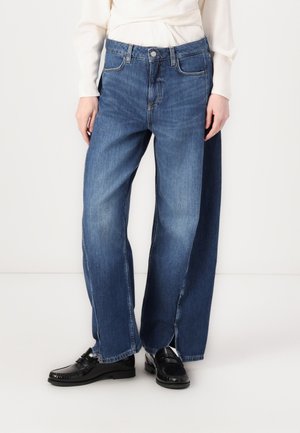 Personne portant un jean en denim bleu à jambes larges avec bouton et fermeture éclair à l'avant, associé à des mocassins noirs et un haut blanc à manches longues.