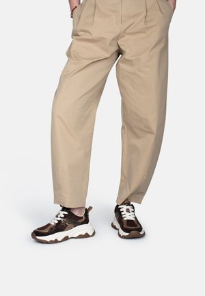 Person wearing beige pleated pants and brown and white chunky sneakers, standing with hands in pockets on white background.