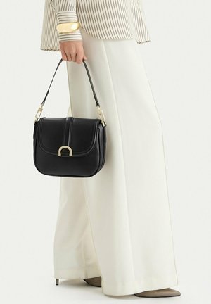 Donna che tiene una borsa nera in pelle con fibbia dorata, indossa pantaloni bianchi a gamba larga e una camicia a righe con braccialetto dorato sul polso.