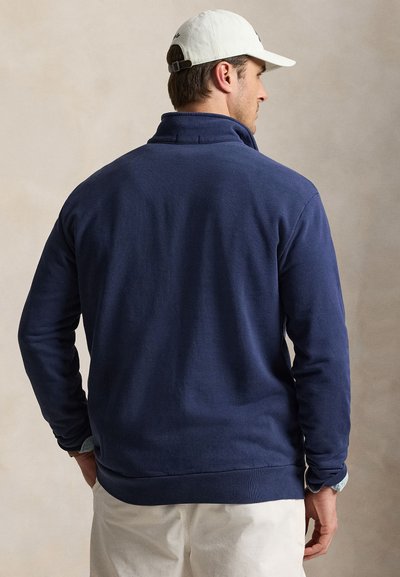 Άνδρας με την πλάτη, φοράει μπλε σκούρο φλις μπουφάν, λευκό καπέλο και ανοιχτόχρωμο παντελόνι μπροστά σε απλό μπεζ φόντο.