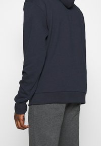 Sudadera azul marino con mangas largas, un acabado texturizado y capucha con cordón. Se combina con pantalones de chándal grises, que muestran un ajuste relajado.
