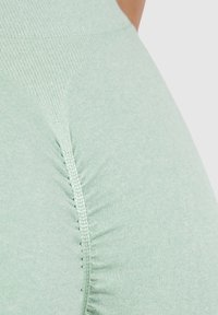 Světlé zelené legíny s texturovanou látkou a nařaseným šitím po stranách, které zdůrazňují pevné uchycení a hladký povrch.