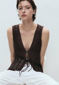 Top tricotat maro cu decolteu în V, având un sistem de închidere cu legătură în față și un design texturat cu fante verticale. Pantaloni albi dedesubt.