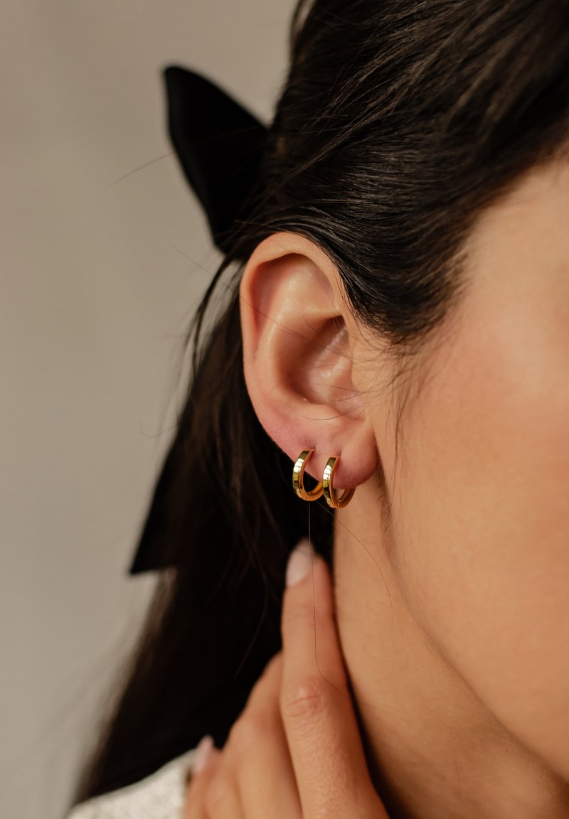 Złote kolczyki w kształcie obrączek z polerowaną powierzchnią, o okrągłym kształcie i gładkiej fakturze, noszone na małżowinie usznej z minimalnymi detalami.