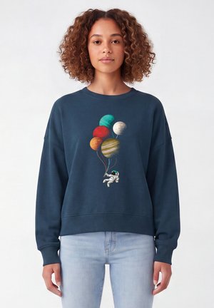 Vrouw draagt een donkerblauwe trui met een astronaut die planeetvormige ballonnen vasthoudt op de voorkant, gecombineerd met lichtblauwe jeans.