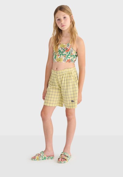 Jeune fille debout portant un crop top fleuri, un short à carreaux jaunes et des sandales assorties à fleurs sur un fond uni.