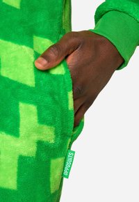 Green fleece onesie with a textured surface, featuring a pixelated pattern. Includes a pocket and ribbed cuffs. Tag reads "OPPOSUITS."