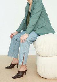 Light green blazer over an olive top, paired with light blue jeans featuring side slits, and dark brown high-heeled mules. Neutral background.