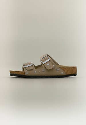 Sandală bej cu două curele reglabile cu cataramă, nituri argintii, talpă interioară din plută și talpă exterioară texturată neagră, prezentată în profil lateral.