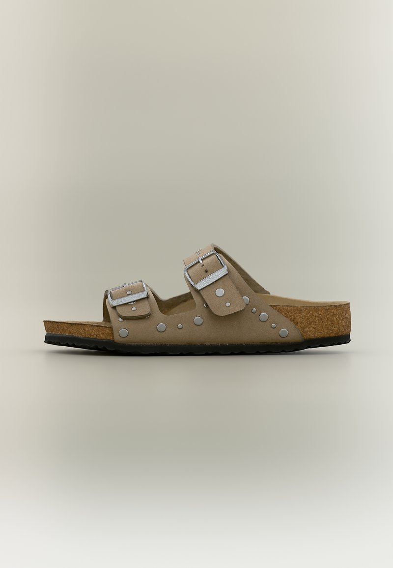 Μπεζ σανδάλι με δύο ρυθμιζόμενους ιμάντες με αγκράφες, ασημένια καρφιά, πάτο από φελλό και μαύρη υφασμάτινη σόλα, εμφανίζεται σε πλάγια όψη.