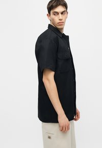 Jeune homme de profil, portant une chemise noire à manches courtes boutonnée et un pantalon beige avec un logo sur la poche, sur un fond uni.