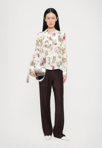 Weiße Bluse mit floralen Mustern in mehreren Farben; plissierte, dunkelbraune weite Hosen; hält eine kleine, runde silberne Tasche; spitze beige Schuhe.