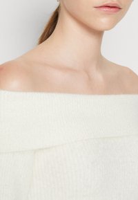 Maglione bianco a spalle scoperte in maglia con un motivo a coste testurizzato. Materiale morbido e leggero, con una scollatura ampia e un design cropped.